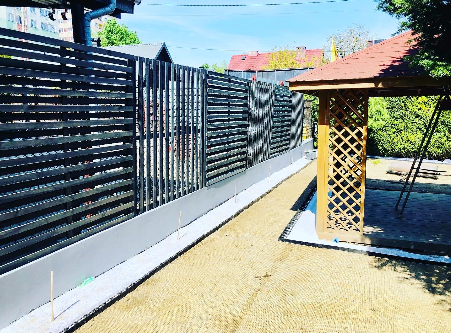 Nowoczesne ogrodzenie z poziomych, ciemnych desek i betonowej podmurówki, z widoczną drewnianą altaną z kratownicą w tle, w słoneczny dzień.