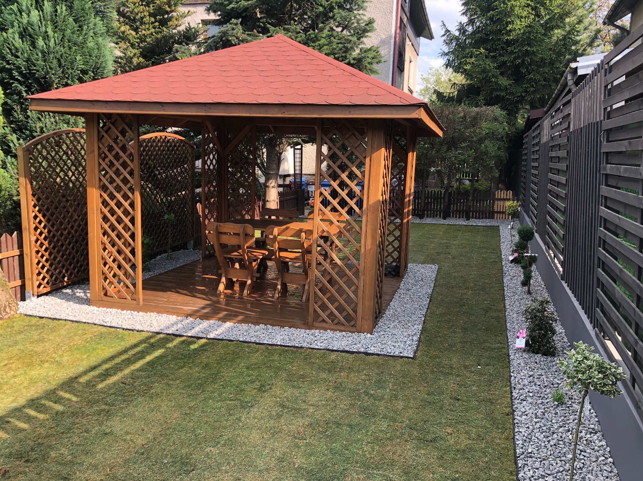 Drewniana altana z czerwonym dachem, otoczona zielonym trawnikiem i szarym żwirem, z widocznym stołem i krzesłami wewnątrz, częściowo zacieniona przez drzewa.