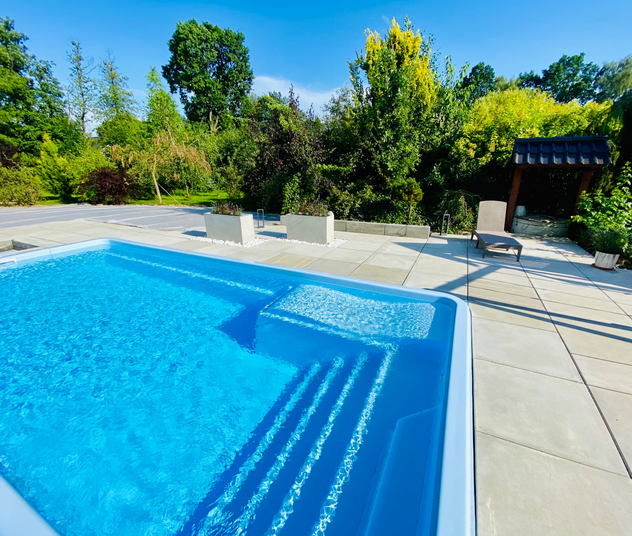 Błękitny basen z wejściem schodkowym, otoczony jasnymi płytami chodnikowymi, donicami z białym kamieniem i zielenią w tle, w słoneczny dzień.