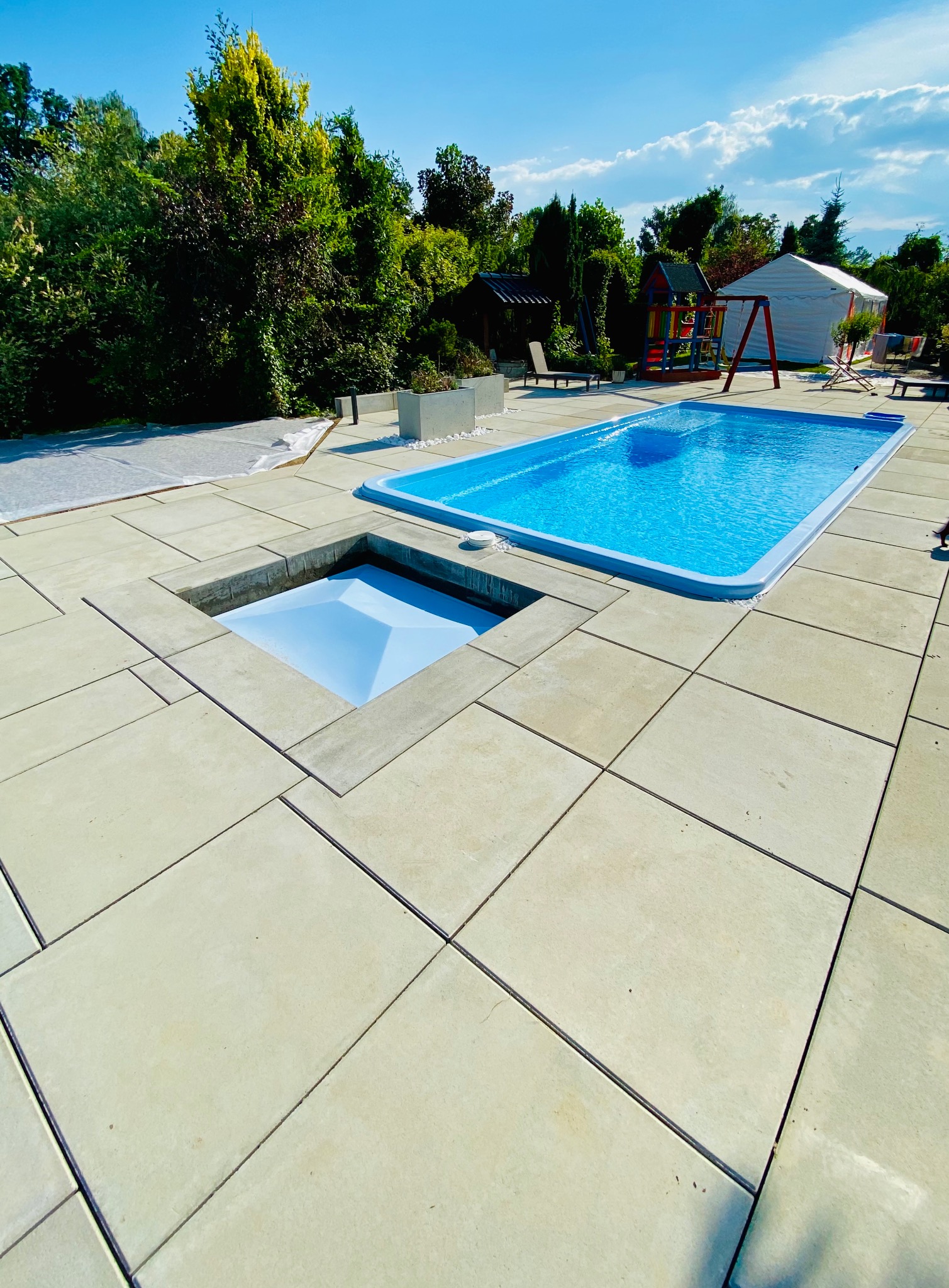 Nowoczesny basen z turkusową wodą i betonową obudową, z ukrytym zejściem, otoczony zielenią i placem zabaw w tle, pod błękitnym niebem.