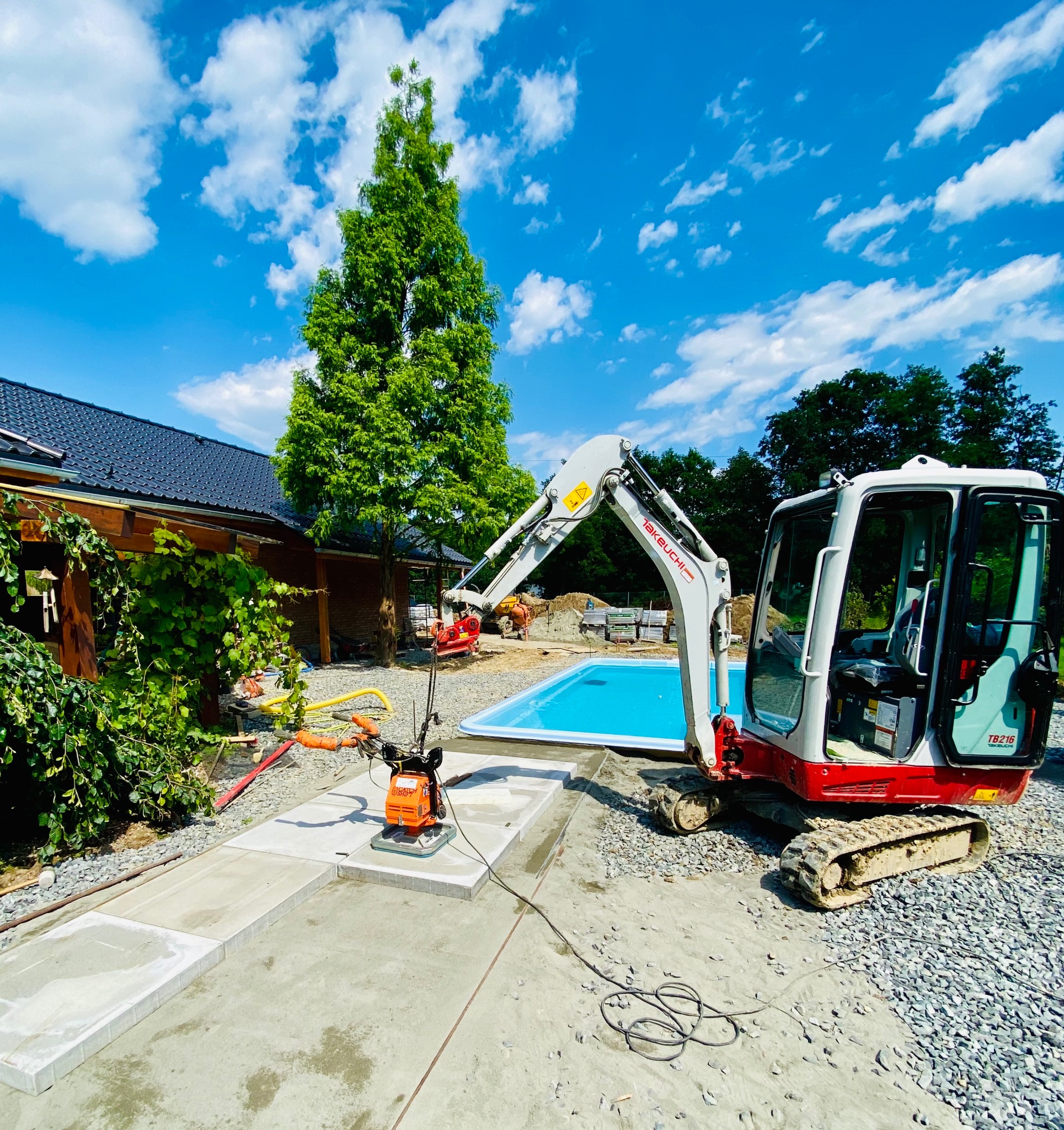 Mini koparka Takeuchi TB216 na żwirowym podłożu obok budowanego basenu z błękitną wodą, w tle budynek z ciemną dachówką i wysokie drzewo pod błękitnym niebem z białymi chmurami.