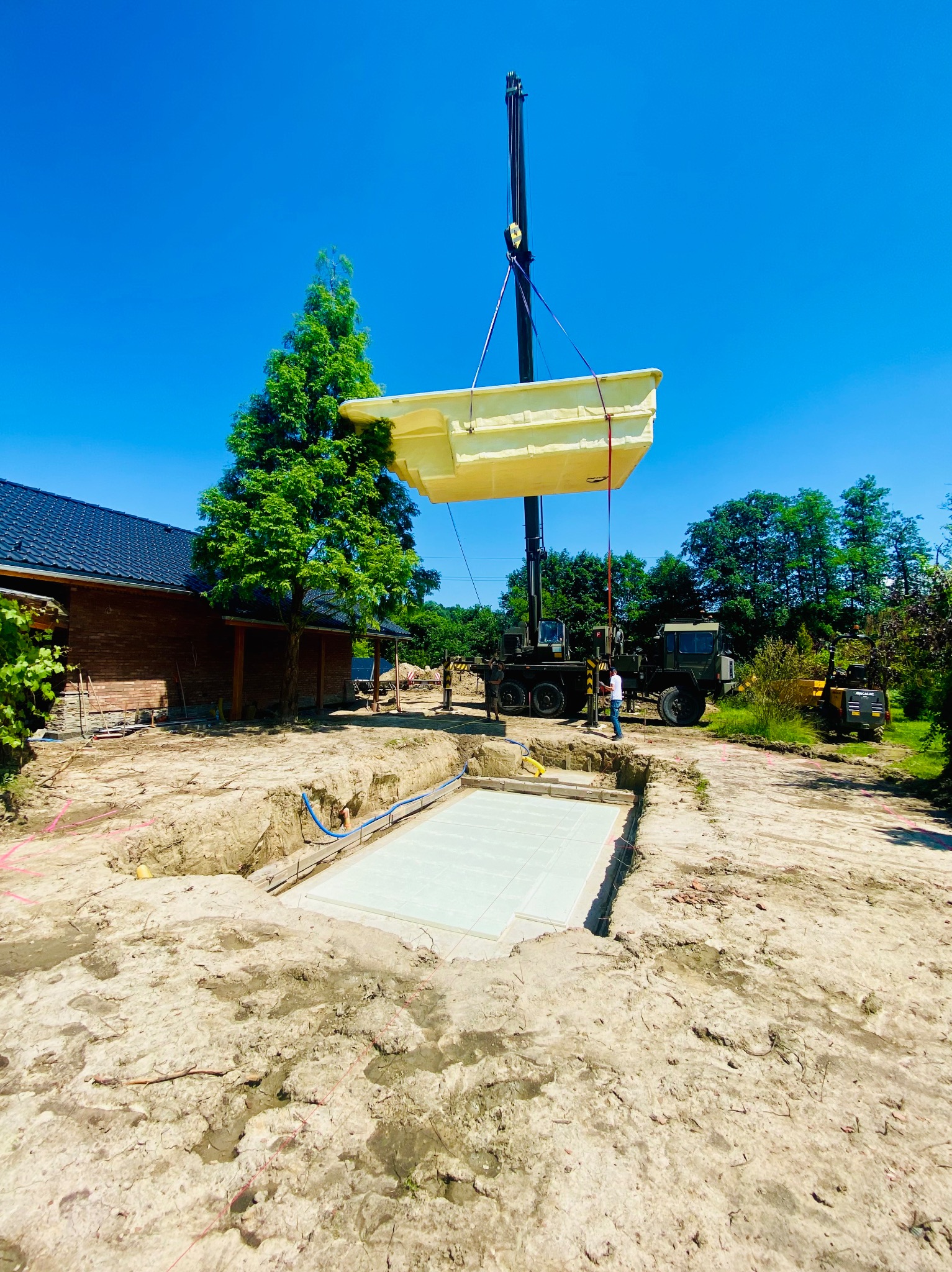 Kran opuszcza prefabrykowany basen do przygotowanego wykopu z betonową płytą fundamentową, widoczny budynek mieszkalny z ciemną dachówką i drzewo iglaste w tle.