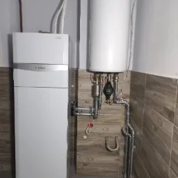 Biała jednostka wewnętrzna z zbiornikiem akumulacyjnym marki Vaillant, widoczne rury i zawory instalacji grzewczej na tle płytek imitujących drewno.