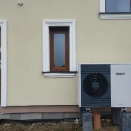 Biało-szara jednostka zewnętrzna pompy ciepła Vaillant aroTHERM, zamontowana na betonowych wspornikach przy ścianie budynku z beżową elewacją, widoczna brązowa stolarka okienna i drzwiowa z białymi...