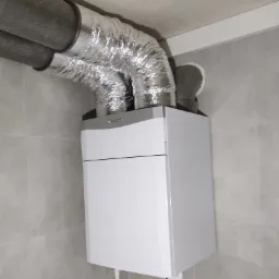 Biały wiszący piec gazowy marki Vaillant z podłączonymi rurami wentylacyjnymi w pomieszczeniu wyłożonym szarymi płytkami.