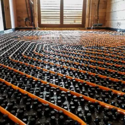 Pomarańczowe rury ogrzewania podłogowego ułożone na czarnych zaczepach, widoczne drewniane ściany i okno w tle.