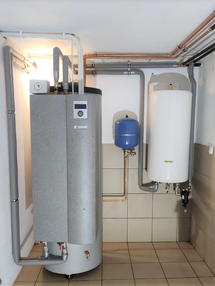 Szary zbiornik buforowy z wyświetlaczem, niebieskie naczynie przeponowe i biały zasobnik ciepłej wody w kotłowni, z widocznymi rurami miedzianymi i szarymi.