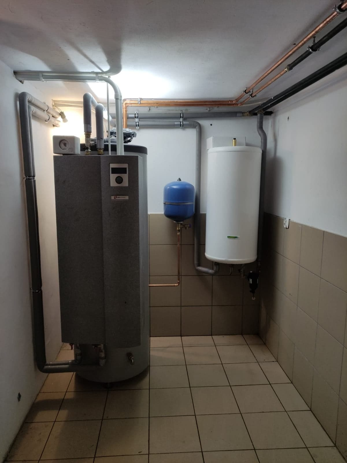 Szara kotłownia z piecem, białym zasobnikiem ciepłej wody i niebieskim naczyniem przeponowym, rury miedziane i PCV pod sufitem, płytki podłogowe w odcieniach beżu.