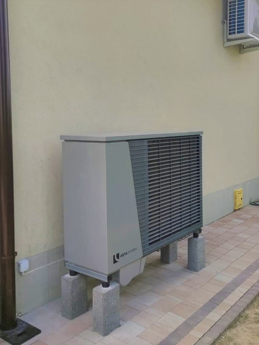 Szara jednostka zewnętrzna urządzenia grzewczego Alpha Innotec, zamontowana na betonowych bloczkach przy beżowej ścianie budynku, z widocznym klimatyzatorem powyżej.
