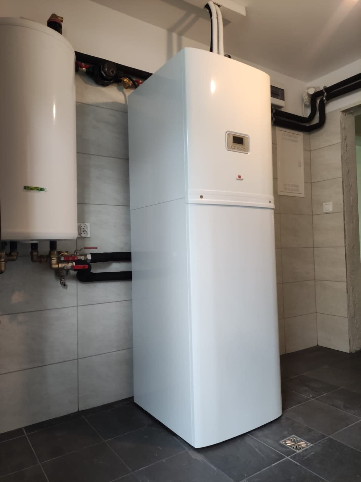 Biała, dwuczęściowa jednostka grzewcza z wyświetlaczem, zainstalowana w pomieszczeniu z szarą glazurą i czarnymi rurami.