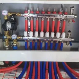 Rozdzielacz ogrzewania podłogowego w skrzynce, widoczne czerwone i niebieskie rury, manometry i pompa obiegowa, ułożone na folii aluminiowej z nadrukowaną siatką.