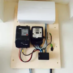 Dwa liczniki elektryczne, czarny i biały, zamontowane na drewnianej płycie, z przewodami i skrzynką rozdzielczą, pod białą obudową instalacyjną na ścianie.