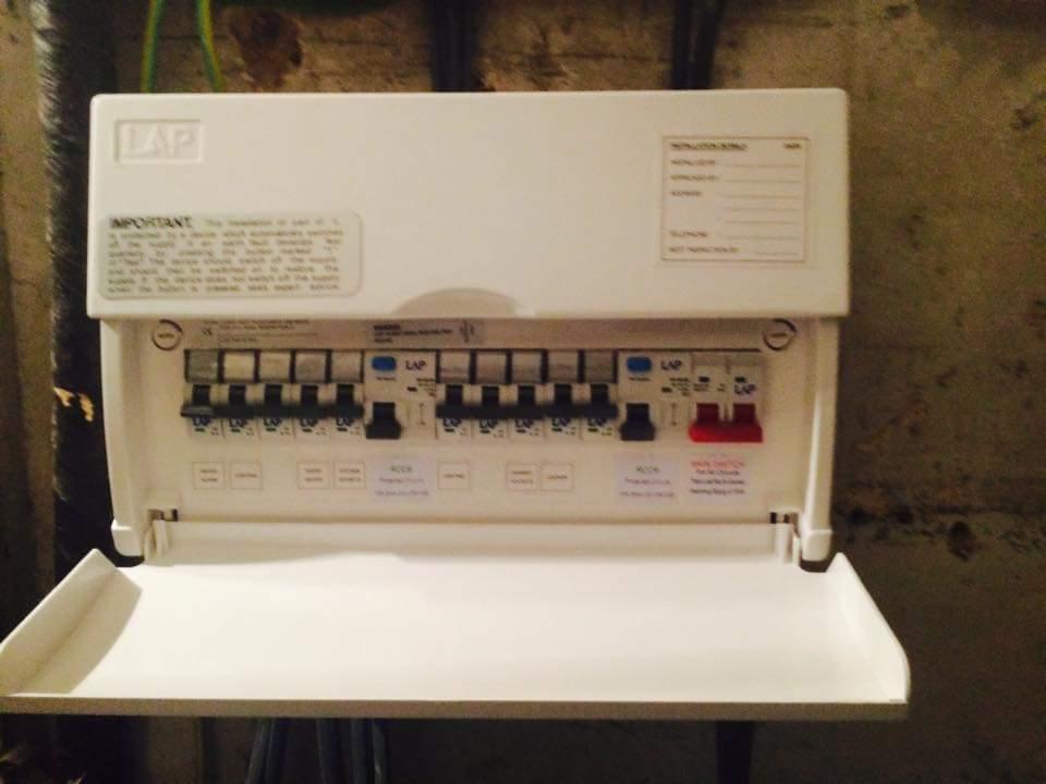 Otwarta, biała skrzynka bezpiecznikowa LAP z widocznymi bezpiecznikami i naklejkami informacyjnymi, zamontowana na szarej, teksturowanej ścianie.