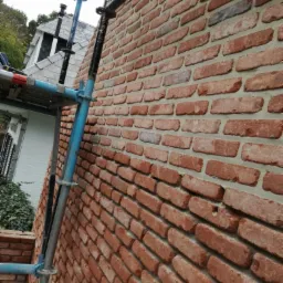 Murowana ściana z czerwonej cegły z widocznym rusztowaniem, w tle fragment budynku z szarą dachówką.