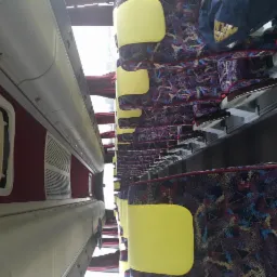 Wnętrze autobusu z rzędami foteli obitych wzorzystą tkaniną, żółte zagłówki, widok z góry na dół, oświetlenie wewnętrzne.