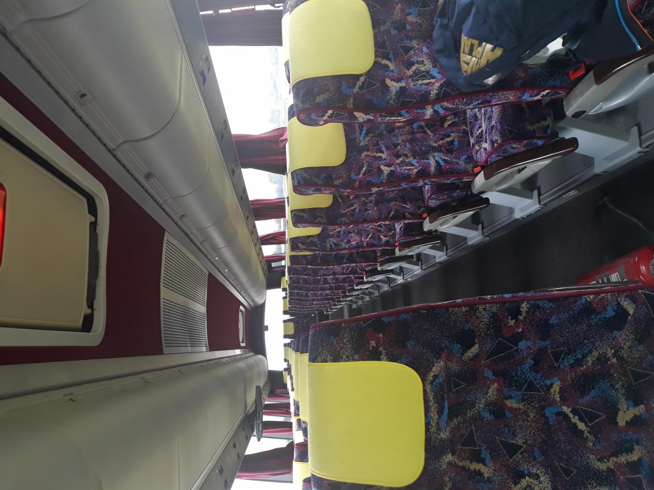 Wnętrze autobusu z rzędami foteli obitych wzorzystą tkaniną, żółte zagłówki, widok z góry na dół, oświetlenie wewnętrzne.