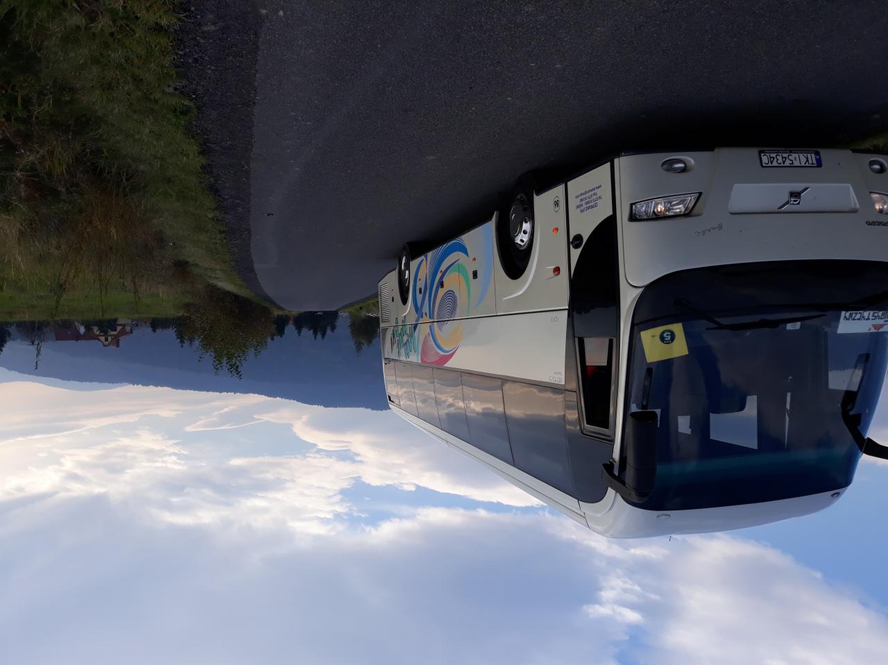 Biały autobus z kolorową grafiką na tle gór i błękitnego nieba z chmurami, stojący na asfaltowej drodze.