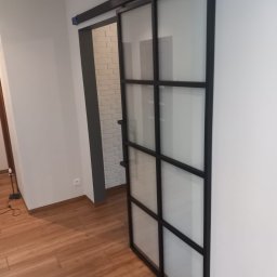 MEDOKIN WELD NIKODEM PLEDZIEWICZ - Przesuwne drzwi szklane z czarną metalową ramą, zamontowane na szynie, widoczne na tle białej ściany i drewnianej podłogi, z fragmentem białej ściany z cegły w tle.