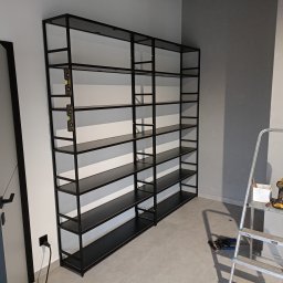 MEDOKIN WELD NIKODEM PLEDZIEWICZ - Czarny, metalowy regał z półkami, ustawiony przy ścianie, poziomica przymocowana do ramy, drabina aluminiowa i narzędzia w pobliżu.