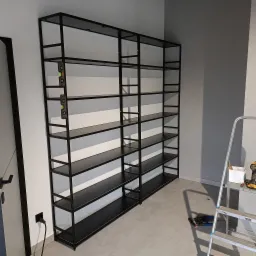 Czarny, metalowy regał z półkami, ustawiony przy ścianie, poziomica przymocowana do ramy, drabina aluminiowa i narzędzia w pobliżu.
