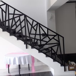 MEDOKIN WELD NIKODEM PLEDZIEWICZ - Nowoczesna, czarna balustrada schodowa o geometrycznym wzorze, widoczna z boku na tle białych ścian i ciemnych stopni, z dekoracyjnym stołem z białym obrusem w dolnej części kadru.