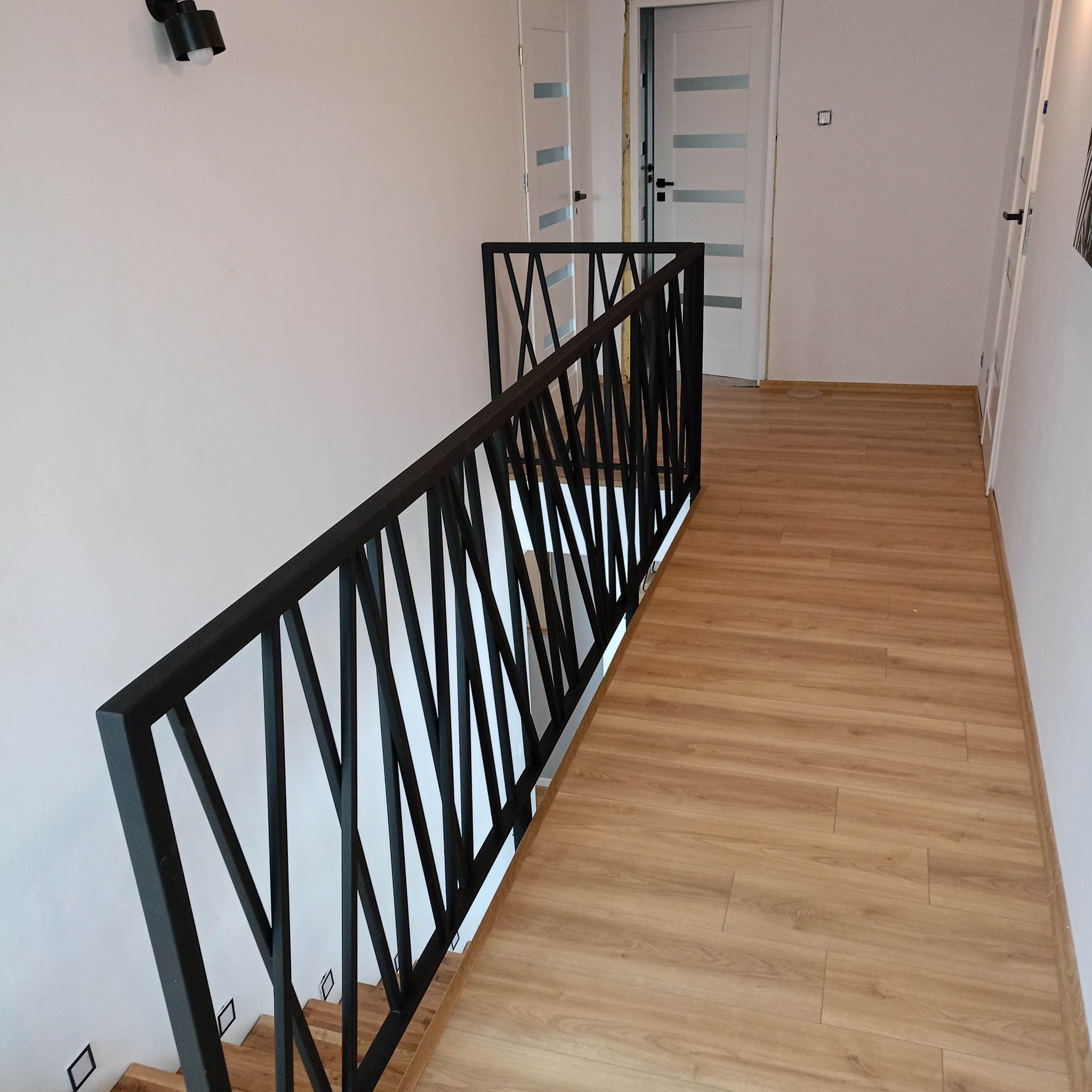 Nowoczesna, czarna barierka schodowa z metalu o geometrycznym wzorze, zamontowana w jasnym wnętrzu z drewnianą podłogą i białymi drzwiami.