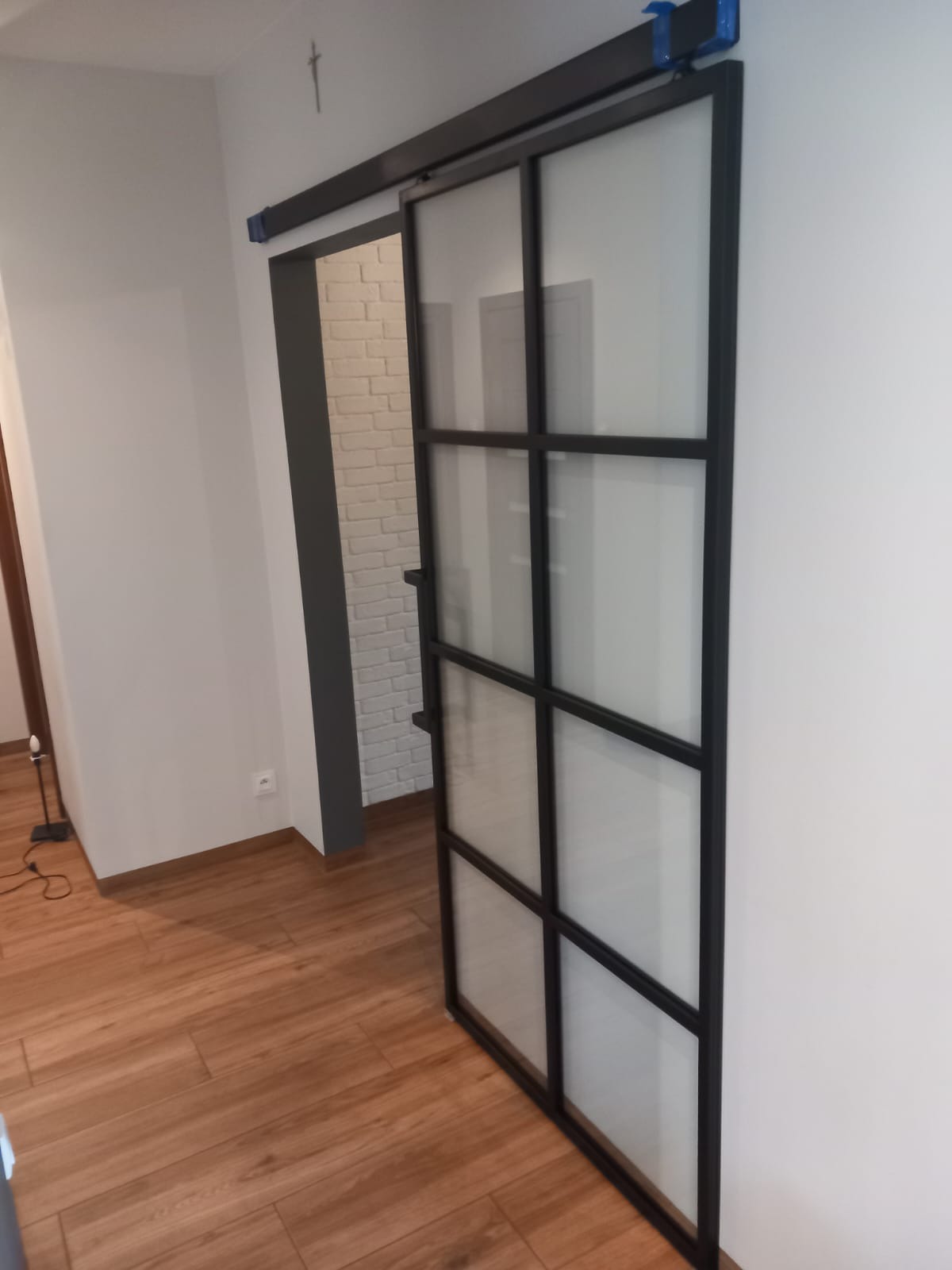 Przesuwne drzwi szklane z czarną metalową ramą, zamontowane na szynie, widoczne na tle białej ściany i drewnianej podłogi, z fragmentem białej ściany z cegły w tle.