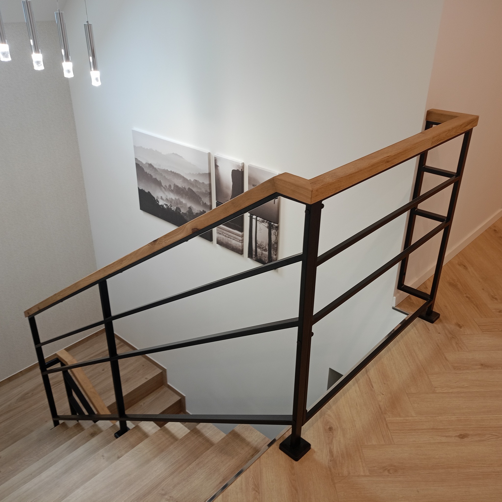 Nowoczesne schody z drewnianymi stopniami i czarną metalową balustradą z drewnianym poręczą, oświetlone wiszącymi lampami i ozdobione czarno-białymi obrazami na ścianie.