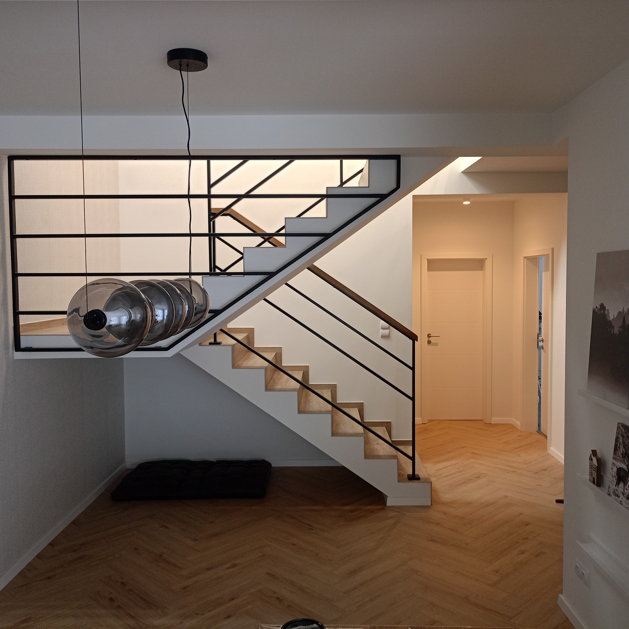 Nowoczesne schody z drewnianymi stopniami i czarną metalową balustradą, uzupełnione wiszącą lampą z kilkoma szklanymi kloszami, na tle parkietu w jodełkę i białych ścian.