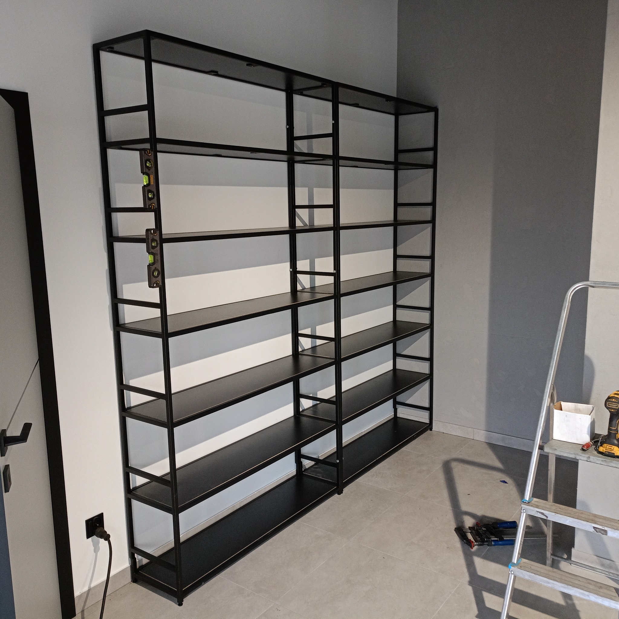 Czarny, metalowy regał z półkami, ustawiony przy ścianie, poziomica przymocowana do ramy, drabina aluminiowa i narzędzia w pobliżu.