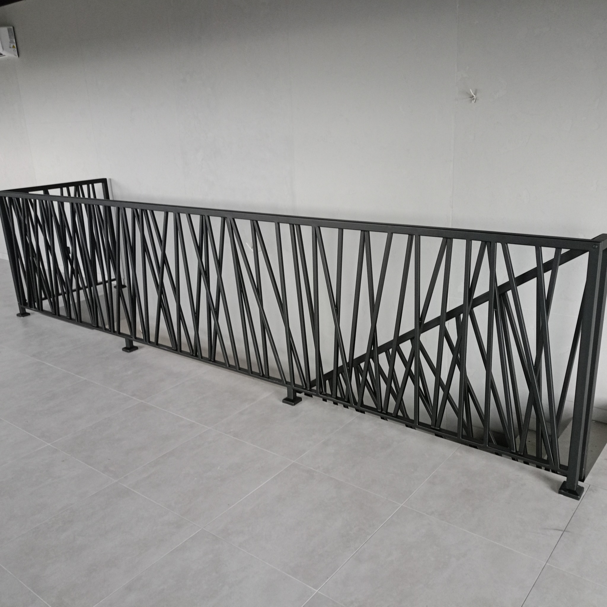 Czarna metalowa barierka o geometrycznym wzorze z ukośnych prętów, zamontowana na jasnoszarej podłodze z płytek, na tle gładkiej, białej ściany. Minimalistyczny design.