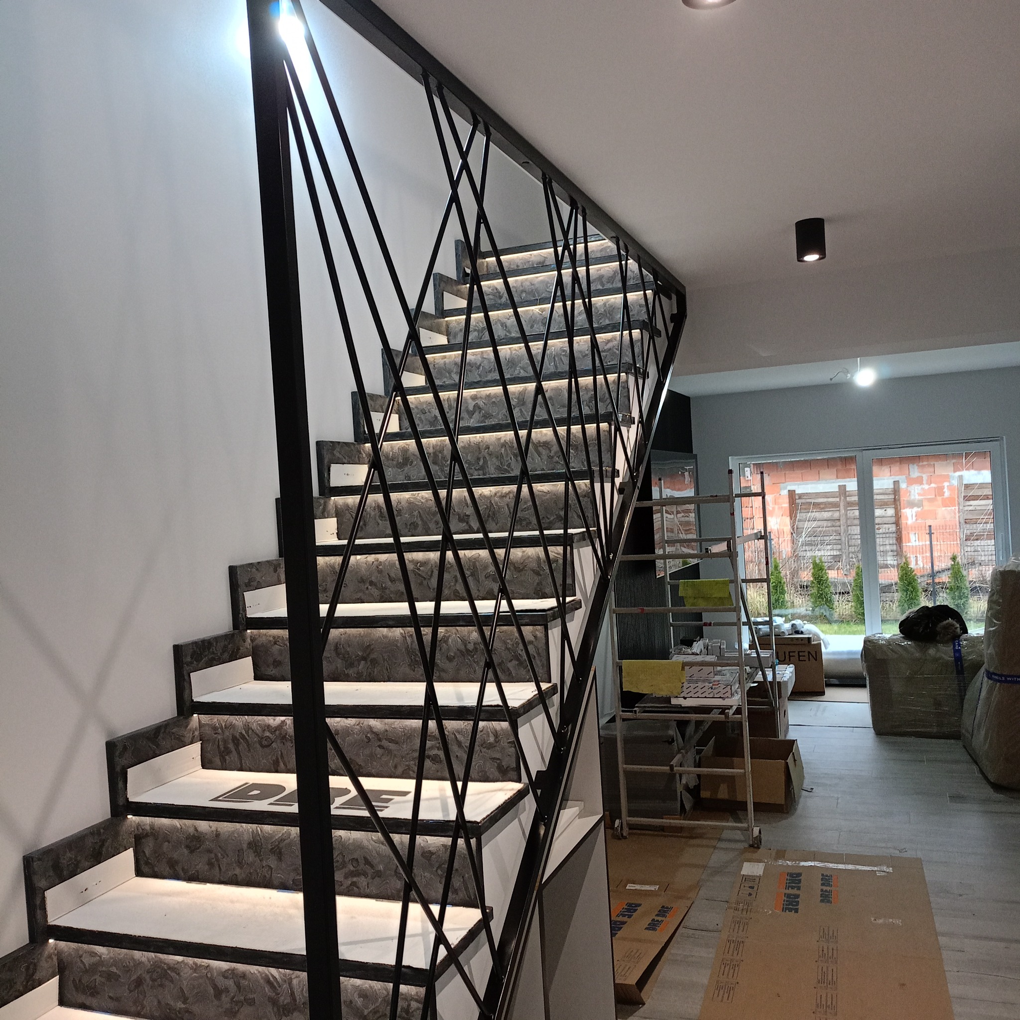 Wewnętrzne schody z podświetlanymi stopniami i metalową balustradą o geometrycznym wzorze, widok z dołu, w tle widoczne kartony i drabina.