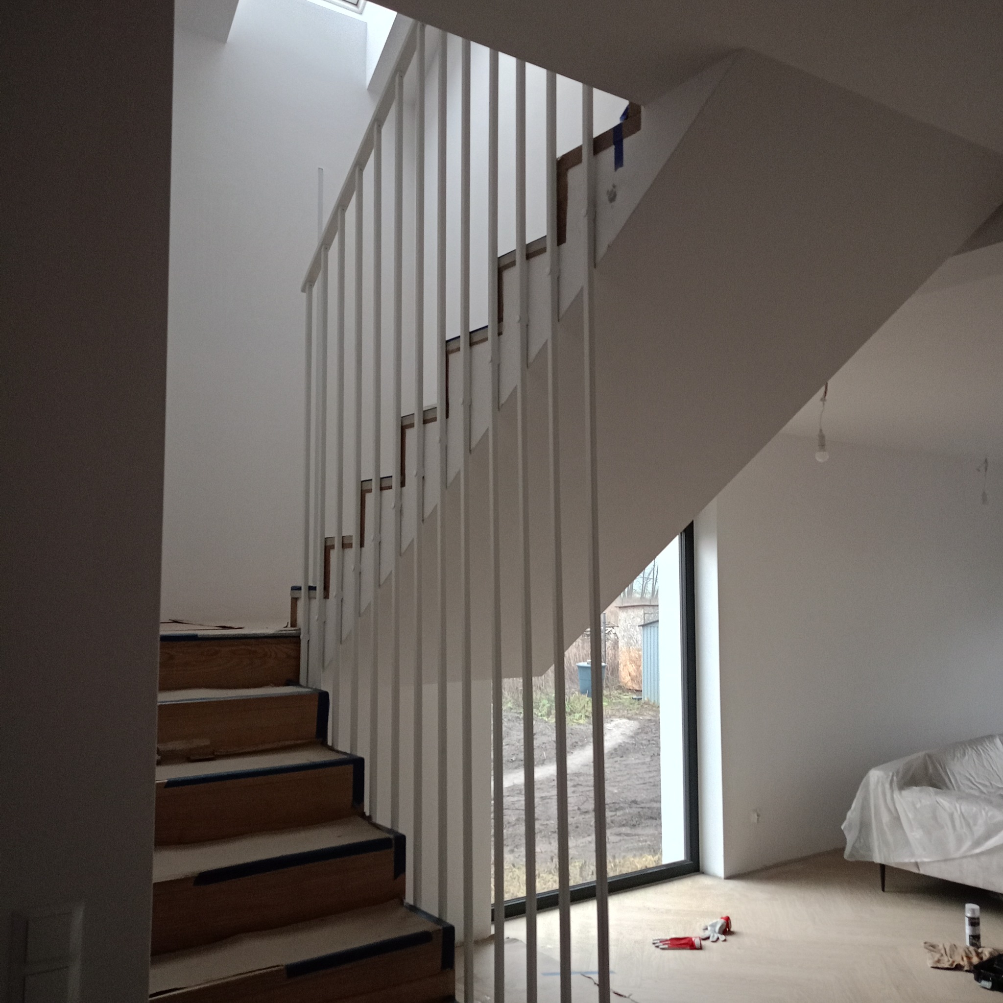 Nowoczesne schody drewniane z białymi, pionowymi balustradami, widok z dołu, fragment okna z widokiem na ogród w tle, pomieszczenie w trakcie wykańczania.