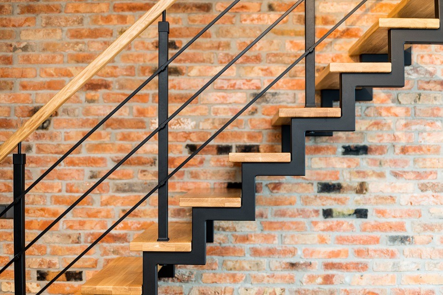 Nowoczesne schody z drewnianymi stopniami i czarną metalową konstrukcją, z poręczą z jasnego drewna, na tle ściany z czerwonej cegły.