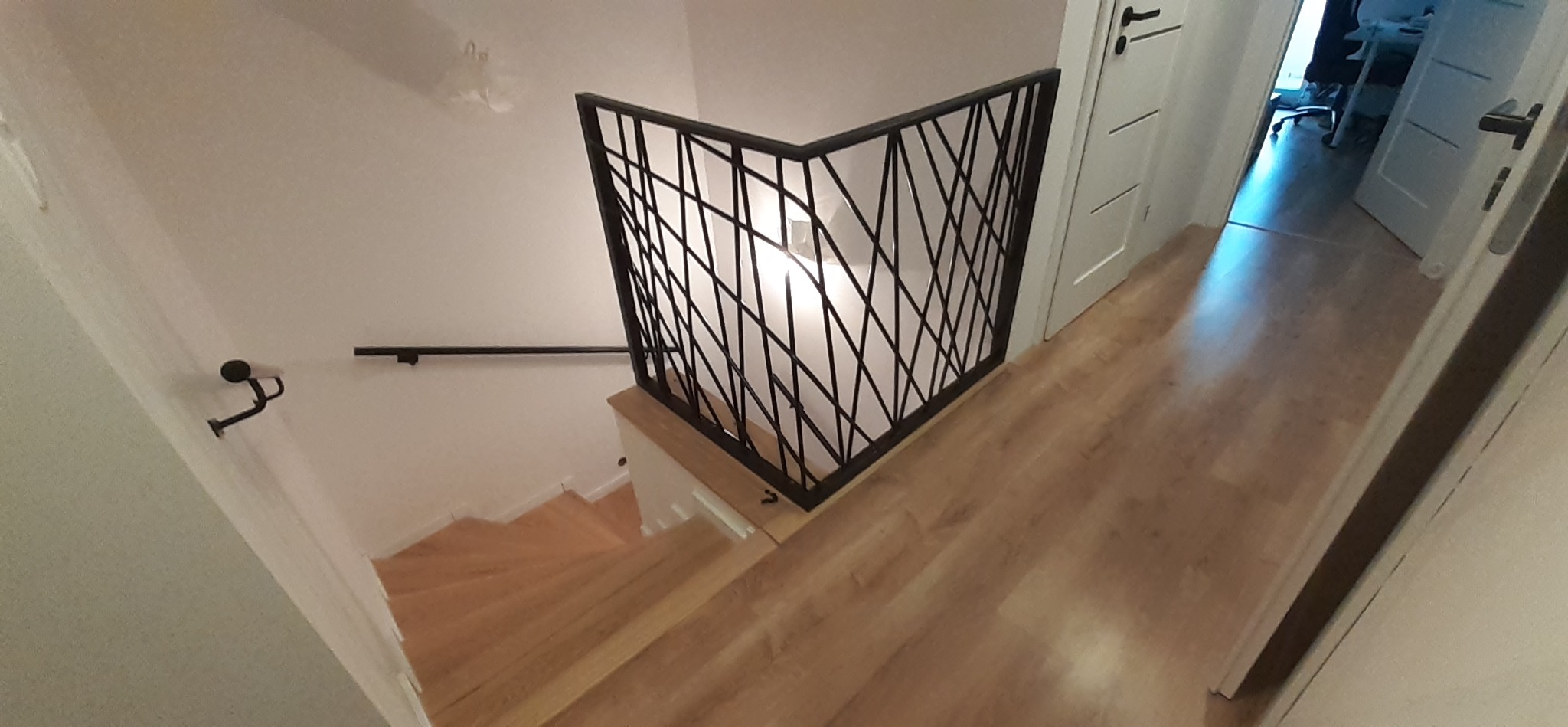 Nowoczesna, czarna, geometryczna barierka schodowa z metalu, widok z góry na drewniane schody i fragment korytarza z białymi drzwiami.