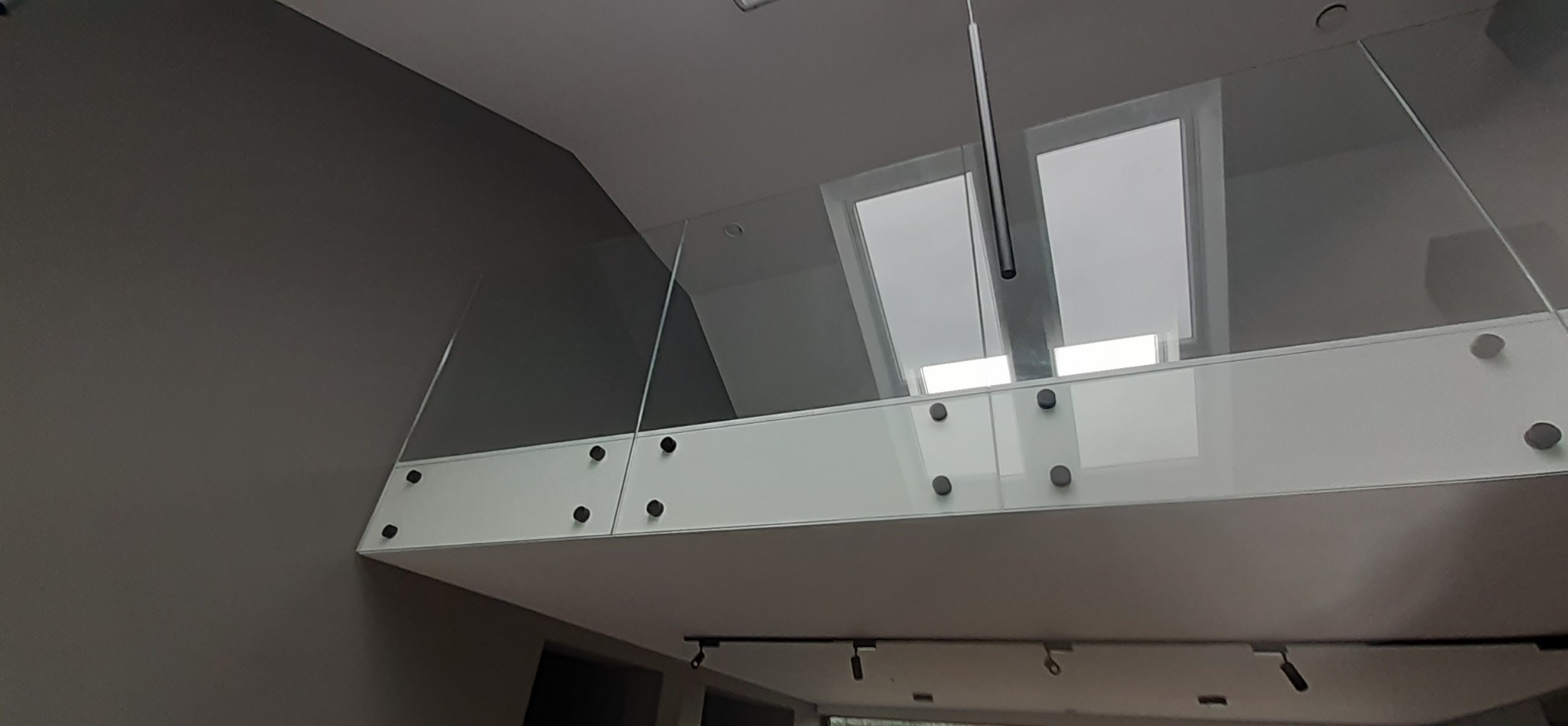 Nowoczesna szklana balustrada z białym panelem i czarnymi mocowaniami, widok z dołu, oświetlenie sufitowe i okna dachowe w tle.