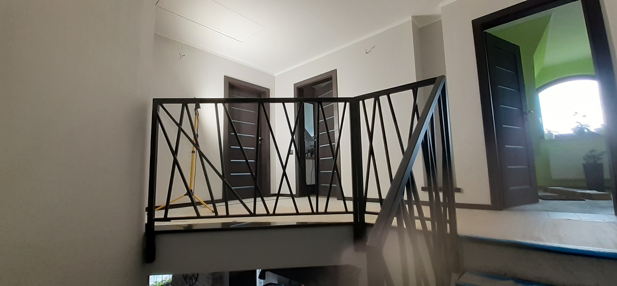 Nowoczesna barierka schodowa z czarnego metalu o geometrycznym wzorze krzyżujących się prętów, widoczna na tle jasnych ścian i ciemnych drzwi wewnętrznych.