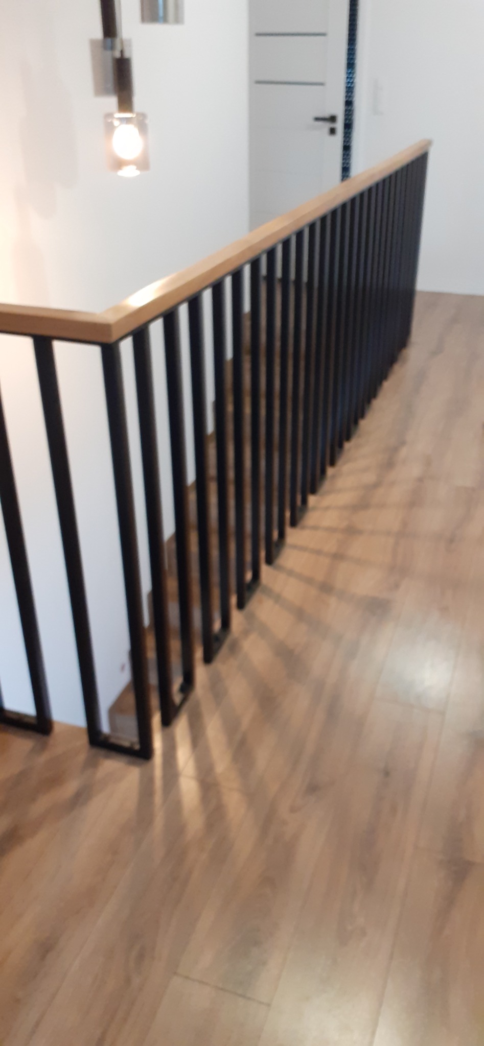 Wewnętrzna barierka schodowa z czarnymi pionowymi prętami i drewnianym blatem, oświetlona lampą ścienną, widok z góry na drewnianą podłogę.