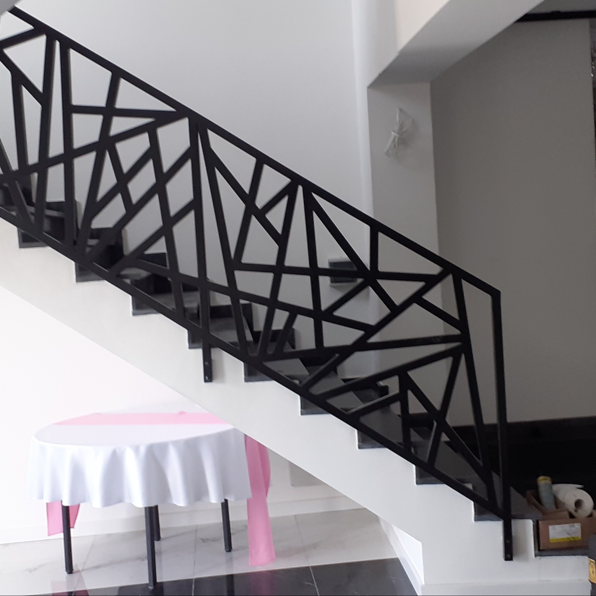 Nowoczesna, czarna balustrada schodowa o geometrycznym wzorze, widoczna z boku na tle białych ścian i ciemnych stopni, z dekoracyjnym stołem z białym obrusem w dolnej części kadru.