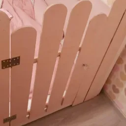 Fragment drewnianej antresoli w pokoju dziecięcym, z widocznymi zawiasami i zabezpieczeniem w kształcie płotku, pomalowanej na jasny różowy kolor, z tapetą w serduszka w tle.