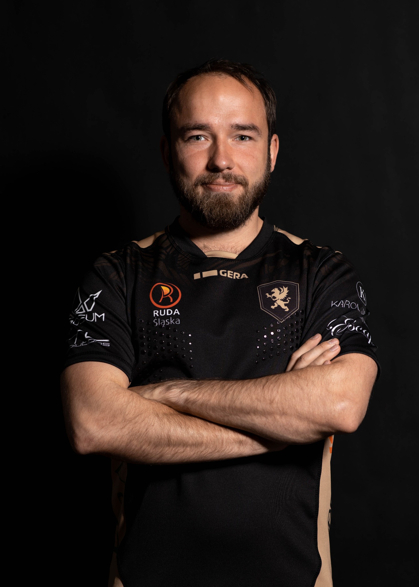 Portret mężczyzny z brodą w czarnej koszulce z logo sponsorów na ciemnym tle, ręce skrzyżowane na piersiach.