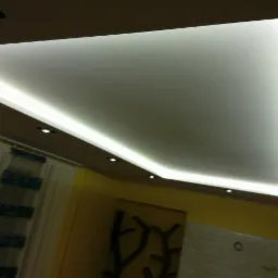 Nowoczesny sufit podwieszany z oświetleniem LED w kształcie litery L, widoczne punktowe lampy halogenowe i fragment zasłon w tle.