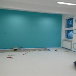 Bauroof Grzegorz Szewczak - Świeżo pomalowane na turkusowo pomieszczenie biurowe z białymi ścianami i wzorami w kółka, widoczne narzędzia malarskie i zabezpieczenia podłogi.