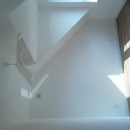 Białe wnętrze poddasza z drewnianą podłogą, ukośnymi ścianami i lampą wiszącą zakrytą folią ochronną, oświetlone promieniami słońca wpadającymi przez okno dachowe.