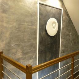 Bauroof Grzegorz Szewczak - Ściana wewnątrz klatki schodowej z dekoracyjną szarą strukturą, obok prostokątna wnęka w ciemniejszym odcieniu z okrągłą, białą lampą, widoczna drewniana balustrada z metalowymi elementami.