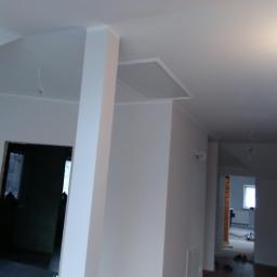 Bauroof Grzegorz Szewczak - Świeżo pomalowane na biało wnętrze mieszkania z widoczną kolumną, sufitem podwieszanym i otwartym przejściem do kolejnego pomieszczenia w trakcie wykańczania.