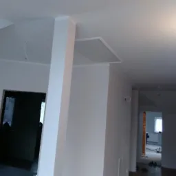 Świeżo pomalowane na biało wnętrze mieszkania z widoczną kolumną, sufitem podwieszanym i otwartym przejściem do kolejnego pomieszczenia w trakcie wykańczania.