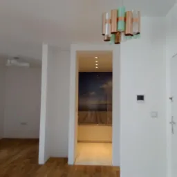 Wnętrze w stanie deweloperskim z drewnianą podłogą, białymi ścianami i dekoracyjną lampą wiszącą w odcieniach miedzi i zieleni, widok na pomieszczenie z fototapetą o tematyce morskiej.