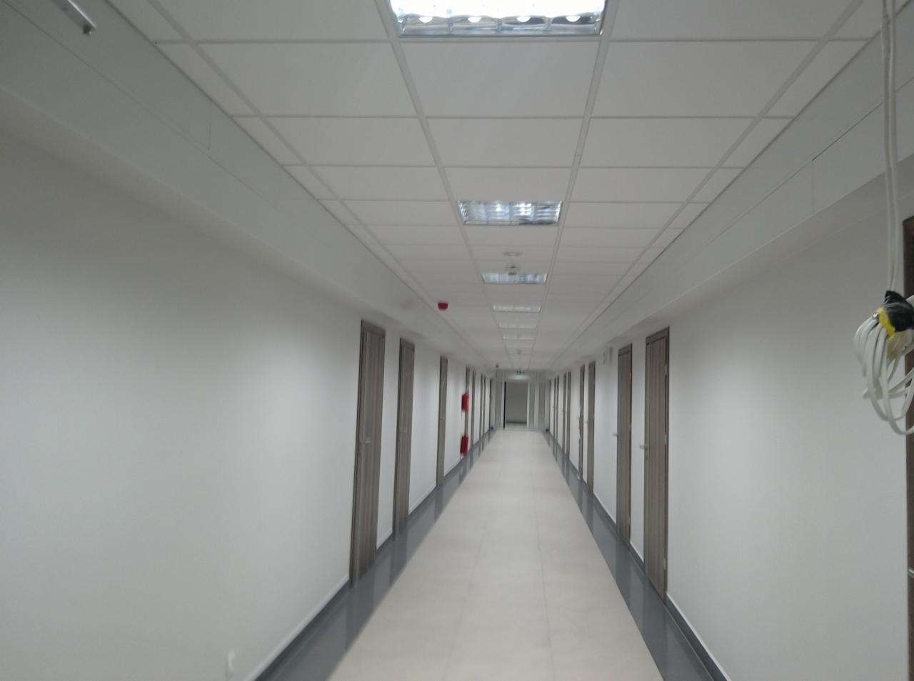 Długi korytarz biurowy z białymi ścianami, szarymi drzwiami i podwieszanym sufitem z lampami rastrowymi, lekko zniekształcony perspektywą.