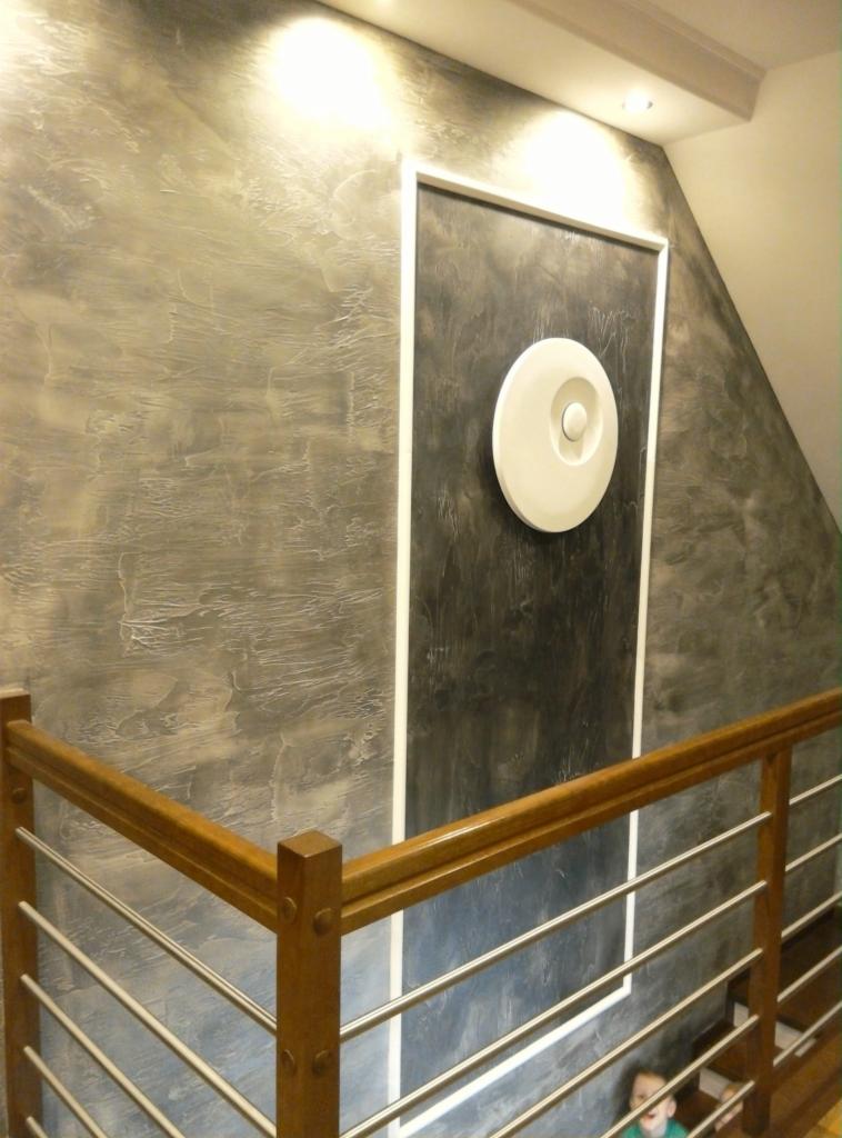 Ściana wewnątrz klatki schodowej z dekoracyjną szarą strukturą, obok prostokątna wnęka w ciemniejszym odcieniu z okrągłą, białą lampą, widoczna drewniana balustrada z metalowymi elementami.